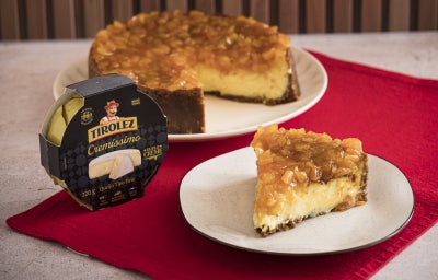 Foto da Receita de Cheesecake de Queijo Brie com Calda de Damasco e Laranja, com uma fatia em um prato à frente, a cheesecake cortada ao fundo e uma embalagem de Queijo Brie Tirolez do lado esquerdo