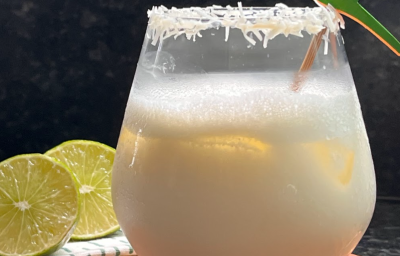 Sobre uma bancada de madeira, um copo transparente com um líquido branco dentro e um pouco de espuma. O copo está decorado com as bordas de coco ralado e um mexedor em formato de papagaio. Ao lado do copo, duas rodelas de limão.