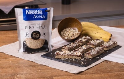 Foto da receita de barrinha proteica de aveia servida em diversas porções sobre uma pedra preta em cima de uma mesa de madeira com um cacho de banana ao fundo Foto da receita de barrinha proteica de aveia servida em diversas porções sobre uma pedra preta em cima de uma mesa de madeira com um cacho de banana ao fundo