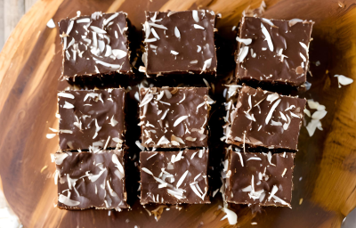 A foto mostra quadradinhos de cocada de chocolate densos e brilhantes em uma travessa rústica de madeira. A cocada tem uma cor marrom profunda, com pedaços de coco visíveis. A foto mostra quadradinhos de cocada de chocolate densos e brilhantes em uma travessa rústica de madeira. A cocada tem uma cor marrom profunda, com pedaços de coco visíveis.