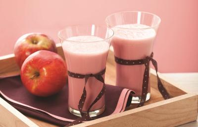 Fotografia em tons de rosa e vermelho, ao centro bandeja de madeira contendo dois copos com creme rosa e decorados com fitas marrom, duas maçãs, sobre guardanapo marrom com borda rosa, tudo sobre bancada de madeira em pátina e fundo rosa.