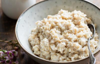 Fotografia em tons claros de um recipiente de vidro branco com acaçá cremoso, e no mesmo recipiente, uma colher de sopa apoiada dentro do acaçá. Ao fundo, molho em um recipiente pequeno na cor branca, sobre uma mesa de madeira na cor marrom escuro.