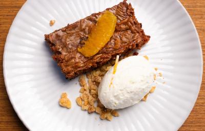 Fotografia de um prato branco raso com um pedaço de bolo de chocolate cremoso com crocante de nozes, e por cima, chantilly de laranja. O prato está sobre uma bancada de madeira clara. Fotografia de um prato branco raso com um pedaço de bolo de chocolate cremoso com crocante de nozes, e por cima, chantilly de laranja. O prato está sobre uma bancada de madeira clara.
