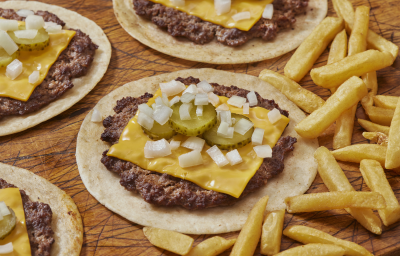Fotografia mostra uma porção de tacos abertos, sob uma mesa de madeira, com recheio de carne bovina, queijo, cebola, alface e picles para finalizar. Fotografia mostra uma porção de tacos abertos, sob uma mesa de madeira, com recheio de carne bovina, queijo, cebola, alface e picles para finalizar.