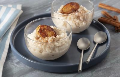 Fotografia em tons de cinza, em uma bancada de madeira escura com um pano azul listrado. Ao centro, um prato redondo preto, com duas taças de arroz doce com banana caramelizada e ao fundo, algumas canelas em pau espalhadas. Fotografia em tons de cinza, em uma bancada de madeira escura com um pano azul listrado. Ao centro, um prato redondo preto, com duas taças de arroz doce com banana caramelizada e ao fundo, algumas canelas em pau espalhadas.