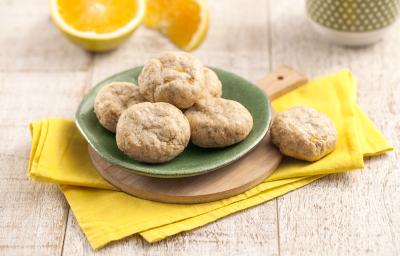 Fotografia em tons de amarelo em uma bancada de madeira clara, um pano amarelo, uma tábua de madeira com um prato verde redondo com alguns biscoitinhos de banana com laranja em cima dele. Ao fundo, uma laranja cortada ao meio e uma xícara verde. Fotografia em tons de amarelo em uma bancada de madeira clara, um pano amarelo, uma tábua de madeira com um prato verde redondo com alguns biscoitinhos de banana com laranja em cima dele. Ao fundo, uma laranja cortada ao meio e uma xícara verde.