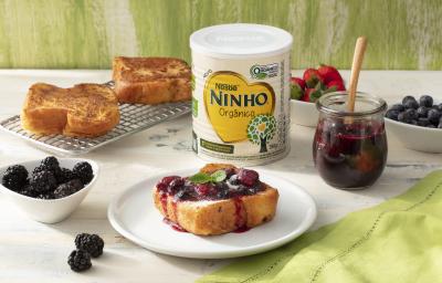 Fotografia em tons de branco e verde de uma bancada branca com um prato branco, sobre ela uma rabanada com geleia. Ao lado um recipientes com frutas vermelhas, uma gradinha com rabanadas, um pote de geleia, uma lata de NINHO orgânico e um paninho verde.