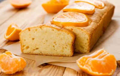 Fotografia de um bolo de mexerica feito em forma pequena e retangular, sobre uma folha de papel manteiga e uma tábua de madeira. Tem uma fatia do bolo cortada na frente, e tem fatias e pedaços de mexerica sobre e ao redor do bolo. Fotografia de um bolo de mexerica feito em forma pequena e retangular, sobre uma folha de papel manteiga e uma tábua de madeira. Tem uma fatia do bolo cortada na frente, e tem fatias e pedaços de mexerica sobre e ao redor do bolo.