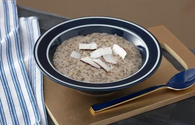 Foto em tons de marrom da receita de mingau de sementes e coco servida em um prato fundo preto com uma colher prateada ao lado sobre uma tábua de madeira lisa com um paninho azul ao lado