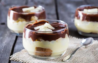 Fotografia em tons de chocolate e branco, com três potinhos contendo um creme branco com biscoitos e coberto com chocolate, um deles sobre toalhinha branca com uma colher ao lado, tudo sobre bancada de madeira em tom roxo.