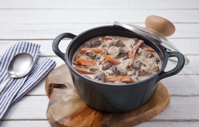 Fotografia em tons de marrom, azul e preto, com panela de ferro preta com tampa ao lado contendo ensopado de carne com legumes e sobre peça de madeira, ao lado guardanapo listrado em branco e azul com um colher por cima, e tudo sobre bancada branca. Fotografia em tons de marrom, azul e preto, com panela de ferro preta com tampa ao lado contendo ensopado de carne com legumes e sobre peça de madeira, ao lado guardanapo listrado em branco e azul com um colher por cima, e tudo sobre bancada branca.