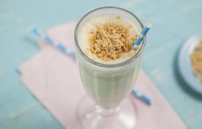 Fotografia em tons de azul e lilás de uma bancada azul com um paninho lilás, sobre ele um copo de milkshake, e dois canudos azuis com bolinhas brancas. Ao lado um prato redondo azul, Fotografia em tons de azul e lilás de uma bancada azul com um paninho lilás, sobre ele um copo de milkshake, e dois canudos azuis com bolinhas brancas. Ao lado um prato redondo azul,
