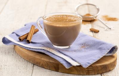 Foto da Receita de Cappuccino Cremoso. Observa-se uma xícara de vidro transparente sobre um guardanapo azul e uma tábua de madeira. Ao fundo, uma peneira com canela em pó. Foto da Receita de Cappuccino Cremoso. Observa-se uma xícara de vidro transparente sobre um guardanapo azul e uma tábua de madeira. Ao fundo, uma peneira com canela em pó.