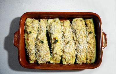 Fotografia em tom claro, com seis abobrinhas em fileira, recheadas e salpicadas com queijo ralado dentro de um refratário retangular marrom escuro, sobre a mesa branca. Fotografia em tom claro, com seis abobrinhas em fileira, recheadas e salpicadas com queijo ralado dentro de um refratário retangular marrom escuro, sobre a mesa branca.