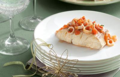 Fotografia em tons de verde em uma bancada de madeira verde, vários pratos brancos redondos empilhados e em cima a posta de peixe com molho de lulas e tomates.
