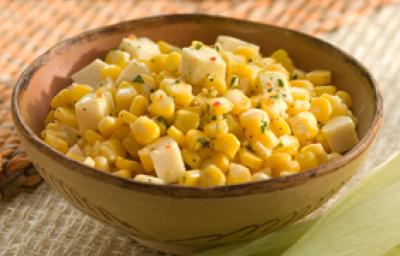 Fotografia em tons de amarelo, bege e rosa, com recipiente em tons de bege contendo milho verde refogado com salsa e pedaços de queijo, ao entorno espigas sobre base de palha marrom , palha de milho, tudo sobre toalha branca Fotografia em tons de amarelo, bege e rosa, com recipiente em tons de bege contendo milho verde refogado com salsa e pedaços de queijo, ao entorno espigas sobre base de palha marrom , palha de milho, tudo sobre toalha branca