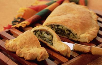 Fotografia em tons de amarelo em uma mesa de madeira com um pano colorido, uma tábua de madeira colorida com o calzone recheado de escarola cortado ao meio