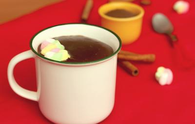 Fotografia em tons de branco e vermelho, ao centro uma xícara branca com chocolate quente e marshmallows, sobre toalha vermelha. Atrás um potinho com achocolatado e uma colher, com paus de canela e marshmallows espalhados ao redor. Fotografia em tons de branco e vermelho, ao centro uma xícara branca com chocolate quente e marshmallows, sobre toalha vermelha. Atrás um potinho com achocolatado e uma colher, com paus de canela e marshmallows espalhados ao redor.