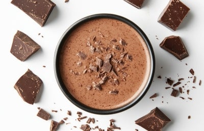 Fotografia vista de cima de uma forma funda, preta, grande e redonda sobre uma mesa branca. Dentro tem a sobremesa, finaliada com a ganache e raspas de chocolate. Ao redor tem pedaços de chocolate. Fotografia vista de cima de uma forma funda, preta, grande e redonda sobre uma mesa branca. Dentro tem a sobremesa, finaliada com a ganache e raspas de chocolate. Ao redor tem pedaços de chocolate.