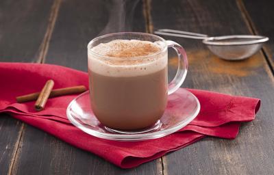 Fotografia em tons de cinza e rosa de uma bancada de madeira escura, um pano rosa escuro com duas canelas em pau, um pires de vidro e sobre ele uma xícara transparente com o cappuccino. Ao fundo uma peneira com canela em pó.