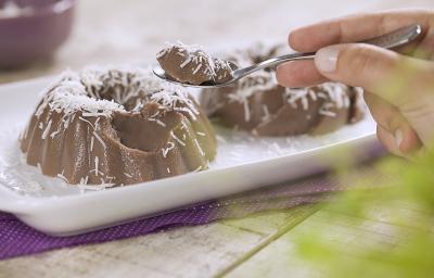 .Fotografia em tons de roxo e branco de uma bancada de madeira branca com um paninho roxo, sobre ele um prato retangular com dois gelados de dark chocolate. Sobre ele uma colher com um pedaço do gelado. Ao fundo um recipiente redondo com coco. .Fotografia em tons de roxo e branco de uma bancada de madeira branca com um paninho roxo, sobre ele um prato retangular com dois gelados de dark chocolate. Sobre ele uma colher com um pedaço do gelado. Ao fundo um recipiente redondo com coco.