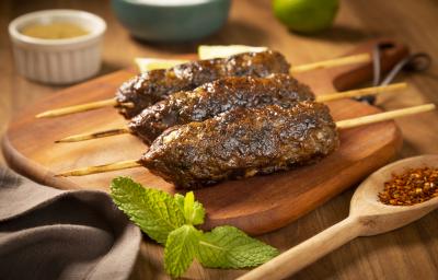 Fotografia em tons de marrom, verde e cinza de uma bancada de madeira marrom vista de frente. Uma tábua de madeira contém 3 espetinhos com carne moída, ao lado uma colher de madeira com tempero, um pano cinza e um potinho redondo branco com tempero. Fotografia em tons de marrom, verde e cinza de uma bancada de madeira marrom vista de frente. Uma tábua de madeira contém 3 espetinhos com carne moída, ao lado uma colher de madeira com tempero, um pano cinza e um potinho redondo branco com tempero.