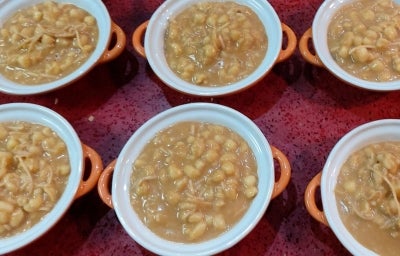 Fotografia de seis mini panelinhas branca e laranja sobre uma toalha de mesa vermelha. Dentro das panelinhas tem canjica com coco e doce de leite.