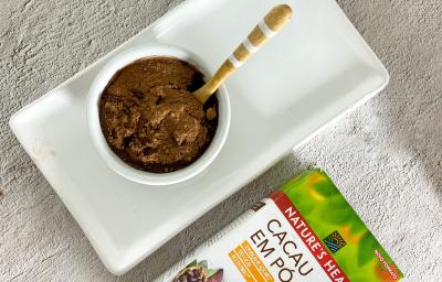 Foto em tons claros de cinza da receita de mousse de cacau com 3 ingredientes servida em um bowl de porcelana branco sobre uma base de porcelana de mesma cor. Ao lado, há uma embalagem de cacau em pó nature's heart Foto em tons claros de cinza da receita de mousse de cacau com 3 ingredientes servida em um bowl de porcelana branco sobre uma base de porcelana de mesma cor. Ao lado, há uma embalagem de cacau em pó nature's heart