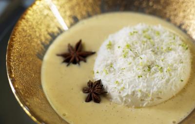 Fotografia de ovos nevados com um creme inglês, algumas raspas de limão por cima e dois anis estrelados no creme. A sobremesa está dentro de um recipiente fundo de alumínio na cor dourado, que está sobre uma mesa preta. Fotografia de ovos nevados com um creme inglês, algumas raspas de limão por cima e dois anis estrelados no creme. A sobremesa está dentro de um recipiente fundo de alumínio na cor dourado, que está sobre uma mesa preta.