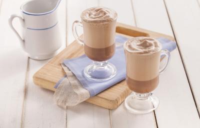 Fotografia em tons de azul em uma bancada de madeira clara, uma tábua de madeira, um paninho azul e duas xícaras de vidro com o cappuccino gelado de cacau dentro deles. Ao fundo, uma jarra branca com azul com mais cappuccino.