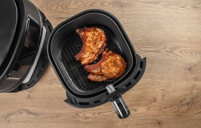 Fotografia vista de cima uma air fryer preta, com a cesta afastada sobre uma mesa de madeira clara. Dentro da Air Fryer tem duas bistecas de porco douradinhas. Fotografia vista de cima uma air fryer preta, com a cesta afastada sobre uma mesa de madeira clara. Dentro da Air Fryer tem duas bistecas de porco douradinhas.