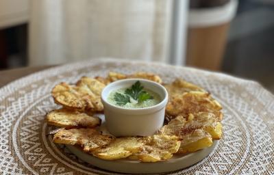 Fotografia em tons de marrom em uma bancada de madeira com uma toalha redonda branca de renda e um prato bege em cima com as batatas assadas e temperadas com um potinho branco ao centro com o molho de maionese verde. Fotografia em tons de marrom em uma bancada de madeira com uma toalha redonda branca de renda e um prato bege em cima com as batatas assadas e temperadas com um potinho branco ao centro com o molho de maionese verde.