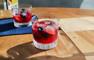 Fotografia de dois copos de vidro baixos com uma bebida sem álcool com frutas vermelhas e gelo. Os copos estão afastados um do outro, e o copo do fundo está sobre um pano azul de tom escuro, sobre uma mesa de madeira. Fotografia de dois copos de vidro baixos com uma bebida sem álcool com frutas vermelhas e gelo. Os copos estão afastados um do outro, e o copo do fundo está sobre um pano azul de tom escuro, sobre uma mesa de madeira.