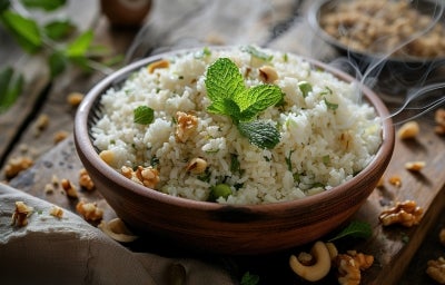 Em uma mesa, tigela de porcelana marrom com porção de arroz branco com castanhas e nozes cortadas em pedaços, decorado com uma folha de hortelã.