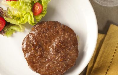 Fotografia em tons de amarelo em uma bancada de madeira, um pano amarelo, um prato redondo branco com salada de alface e tomate e ao lado uma carne de hambúrguer.