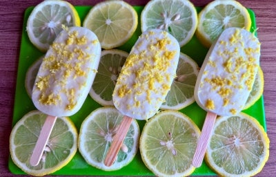 Fotografia em tons claros de verde e amarelo com várias fatias de limão siciliano ao centro. Em cima da cama de limão, existe 3 picolés de limão siciliano cobertos com raspas de limão siciliano Fotografia em tons claros de verde e amarelo com várias fatias de limão siciliano ao centro. Em cima da cama de limão, existe 3 picolés de limão siciliano cobertos com raspas de limão siciliano