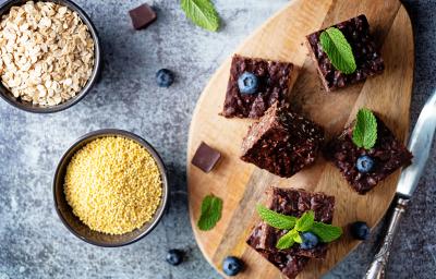 Fotografia em tons de marrom, cinza e amarelo em uma bancada de mármore cinza, uma tábua de madeira e em cima dela 5 brownies de chocolate. Ao lado, potinhos com farinha de aveia e uma faca.