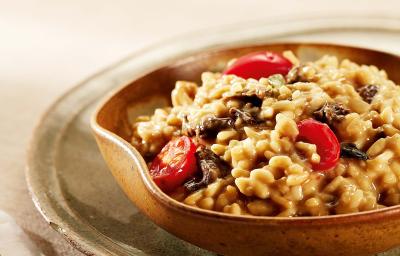 Fotografia em tons de vermelho em uma mesa com uma toalha bege, um prato verde raso grande, um bowl de madeira com o risoto de funghi com tomatinhos dentro dele.