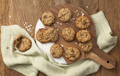 Fotografia em tons de marrom e bege, com tábua de cozinha ao centro com cookies assados e sobre guardanapo bege, tudo em bancada de madeira marrom com flocos de aveia espalhados.