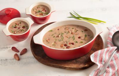 Fotografia em tons de branco e vermelho de uma bancada vista de cima. Em cima de uma tábua redonda contém um recipiente oval com alças nas cores vermelho e branco com a sopa e pedaços de pinhão por cima, ao lado dois potinhos com a sopa e um pano Fotografia em tons de branco e vermelho de uma bancada vista de cima. Em cima de uma tábua redonda contém um recipiente oval com alças nas cores vermelho e branco com a sopa e pedaços de pinhão por cima, ao lado dois potinhos com a sopa e um pano