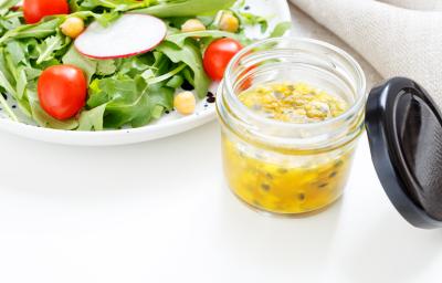 fotografia em tons de branco, verde e amarelo de uma bancada branca vista de cima. Contém um pote transparente com molho de maracujá e ao lado um prato redondo branco com salada.