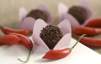 Fotografia em tons de vermelho em uma bancada de madeira com uma toalha branca, ao centro um brigadeiro em uma forminha rosa e várias pimentas vermelhas espalhadas pela mesa.