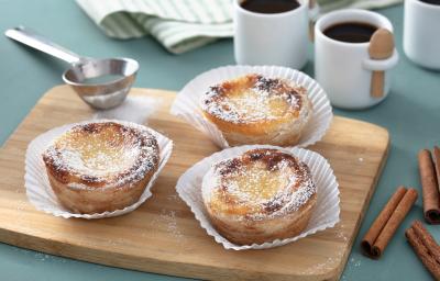 Fotografia em tons de amarelo em uma bancada de madeira de cor azul. Ao centro, uma tábua de madeira contendo os pasteis. Ao lado, há uma peneira de inox e ao fundo, duas xícaras de café. Fotografia em tons de amarelo em uma bancada de madeira de cor azul. Ao centro, uma tábua de madeira contendo os pasteis. Ao lado, há uma peneira de inox e ao fundo, duas xícaras de café.