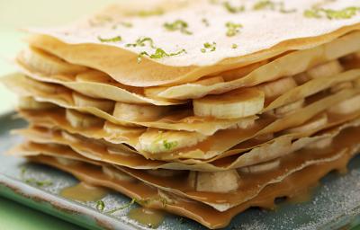Fotografia em tons de verde em uma bancada de madeira com uma toalha verde, um prato quadrado com a torta crocante com recheio de doce de leite e bananas em rodelas. Fotografia em tons de verde em uma bancada de madeira com uma toalha verde, um prato quadrado com a torta crocante com recheio de doce de leite e bananas em rodelas.