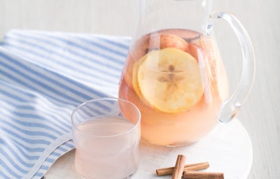 A imagem apresenta um jarro de vidro com um líquido rosa claro, contendo fatias de maçã e paus de canela, sugerindo uma bebida refrescante. Ao lado, há um copo pequeno com a mesma bebida. A cena está sobre uma tábua de mármore branca