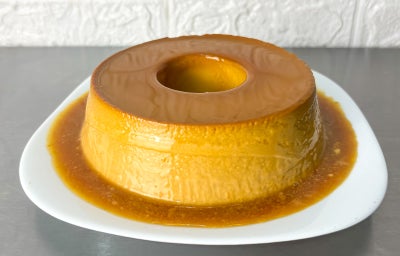 Fotografia em tons claros com um pudim de doce de leite ao centro. O pudim possui uma cor forte de doce de leite e está coberto com uma calda dourada