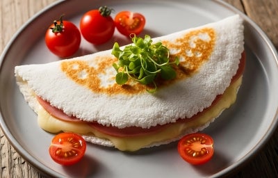 Fotografia em tons claros de uma tapioca recheada com queijo, presunto, orégano e alguns tomatinhos em volta, dentro de um prato cinza de vidro raso. Fotografia em tons claros de uma tapioca recheada com queijo, presunto, orégano e alguns tomatinhos em volta, dentro de um prato cinza de vidro raso.