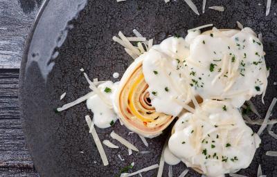 Fotografia vista de cima de três unidades de rondelli deitados em um prato raso escuro. Por cima das massas tem molho branco com queijo parmesão ralado. O prato está sobre uma mesa de madeira preta. Fotografia vista de cima de três unidades de rondelli deitados em um prato raso escuro. Por cima das massas tem molho branco com queijo parmesão ralado. O prato está sobre uma mesa de madeira preta.