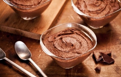Fotografia em tons de marrom com três potes de vidro ao centro. Em cima de cada pote existe uma receita de mousse de chocolate com chia e ninho adulto.