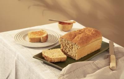 Foto em tons de bege da receita de pão com farinha de aveia servida sobre uma base de porcelana verde-escuro cortada a uma fatia com uma faca de cozinha ao lado. Ao fundo há outra fatia do pão servida em um prato bege claro e um pote de madeira com mel